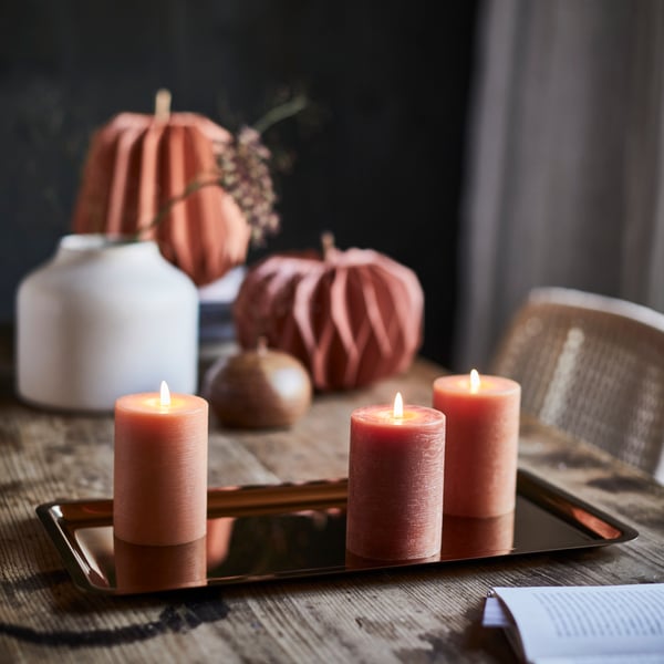Three HÖSTAGILLE unscented pillar candles are placed on a shiny HÖSTAGILLE tray, surrounded by autumn decorations.