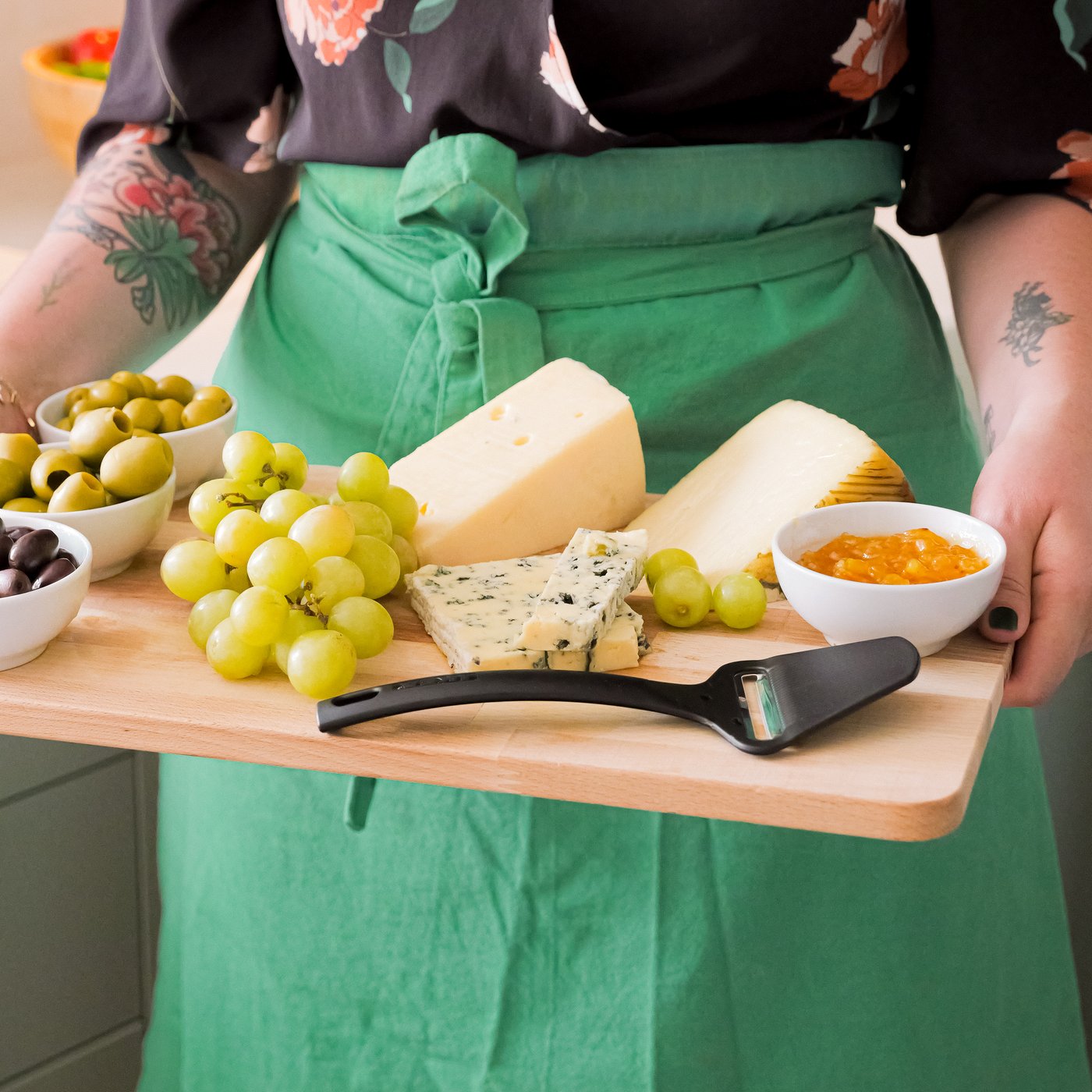 Una donna in grembiule trasporta un tagliere PROPPMÄTT pieno di formaggi, frutta e olive servite nelle ciotole IKEA 365+.