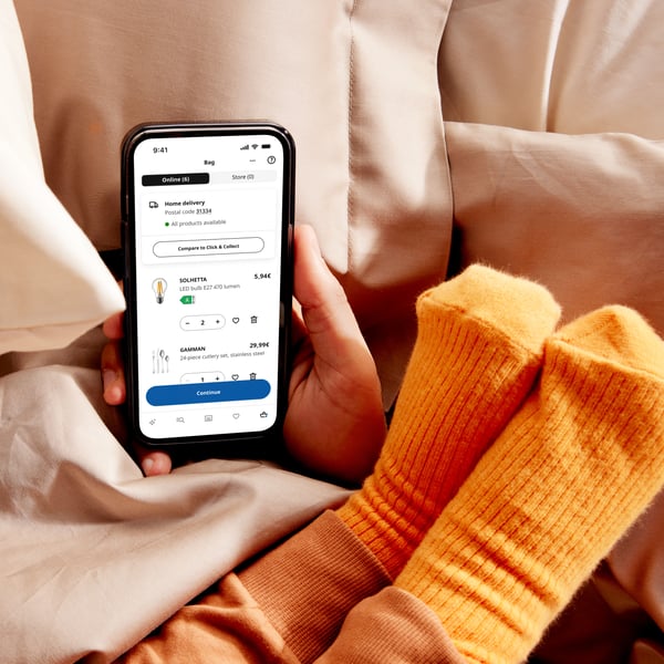 Person nutzt die IKEA App auf dem Smartphone, zeigt Produktdetails und Bewertungen, während sie entspannt auf einem beigen Bett liegt. Daneben orange Socken.
