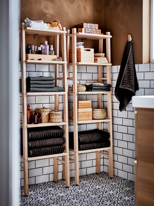 A bright and white-tiled bathroom with a RÅGRUND shelf unit in bamboo stocked with towels, beauty products and boxes.