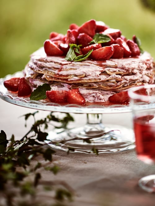 Nahaufnahme einer ARV BRÖLLOP Platte auf Fuß mit Abdeckung, auf der Pfannkuchen mit Erdbeeren liegen. Daneben steht ein IKEA 365+ Kelchglas.