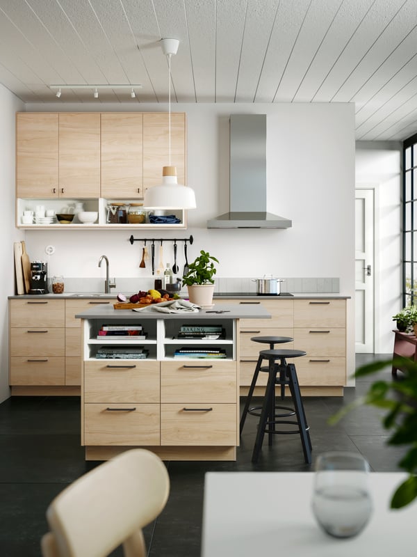 A kitchen island has drawers with FRÖJERED fronts in light bamboo, a big white pendant lamp above it and two black stools.