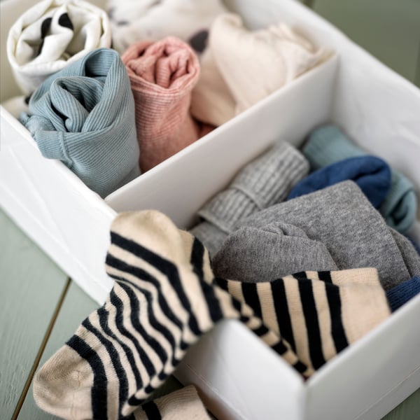 A white STUK box with compartments on a green floor holding different types of rolled clothing and coloured socks.