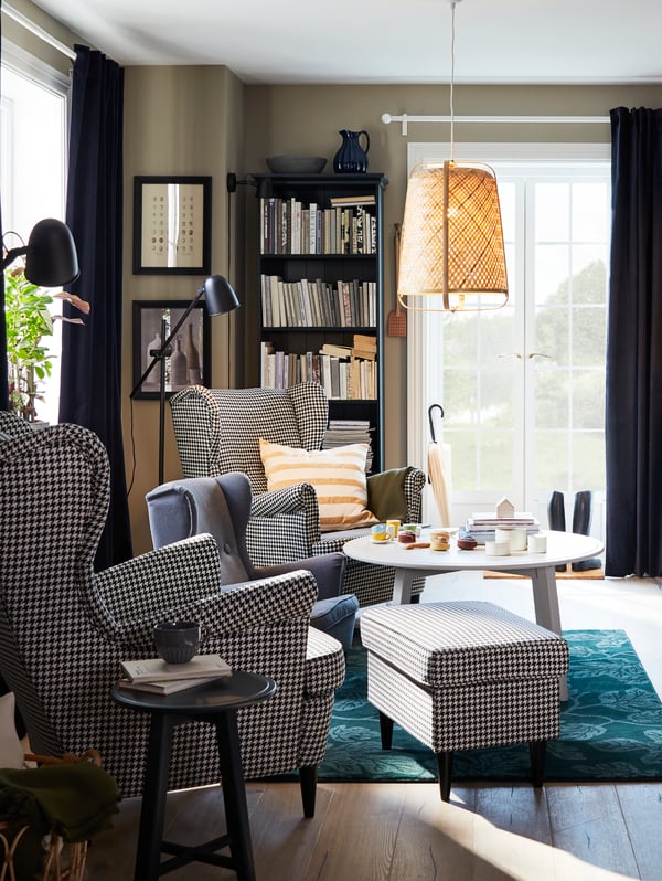 A beige STRANDMON wing chair with a matching footstool, a black KVISTBRO storage table and a reading lamp next to it.
