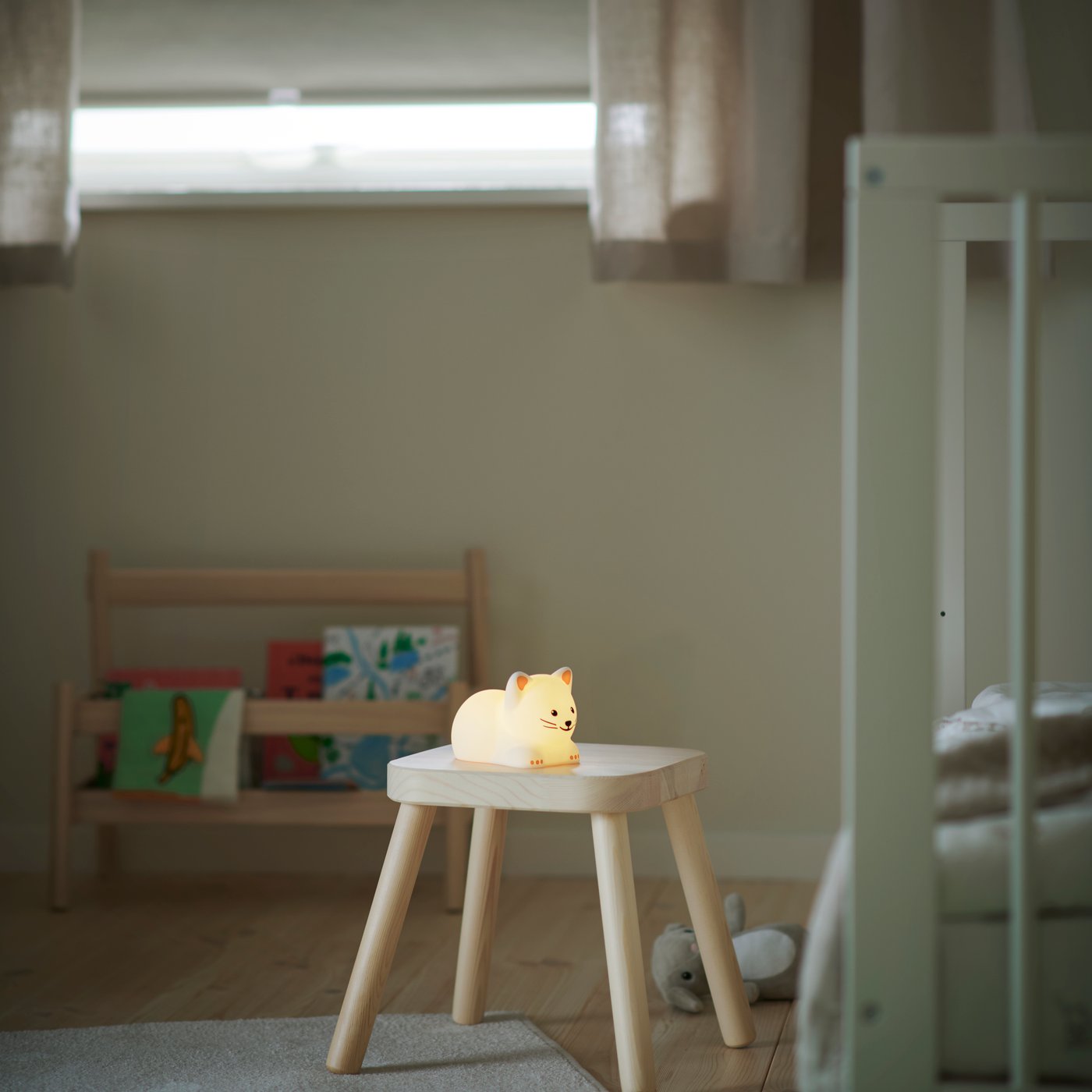 A small wooden stool with a glowing TÖVÄDER LED night light, placed in a neutral children’s room with bookshelves nearby.
