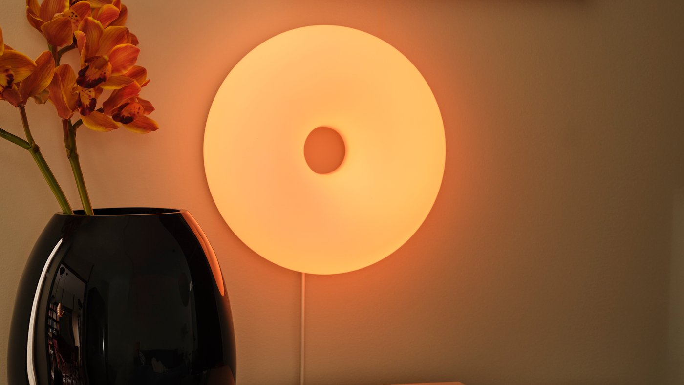 A VARMBLIXT LED table/wall lamp is mounted on the wall above a BESTÅ TV bench. A black vase stands on the TV bench.