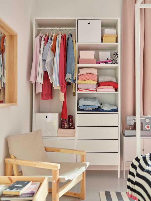 Dans une chambre lumineuse, une armoire-penderie AURDAL blanche avec des vêtements sur une tringle, des boîtes et des vêtements colorés sur des tablettes.