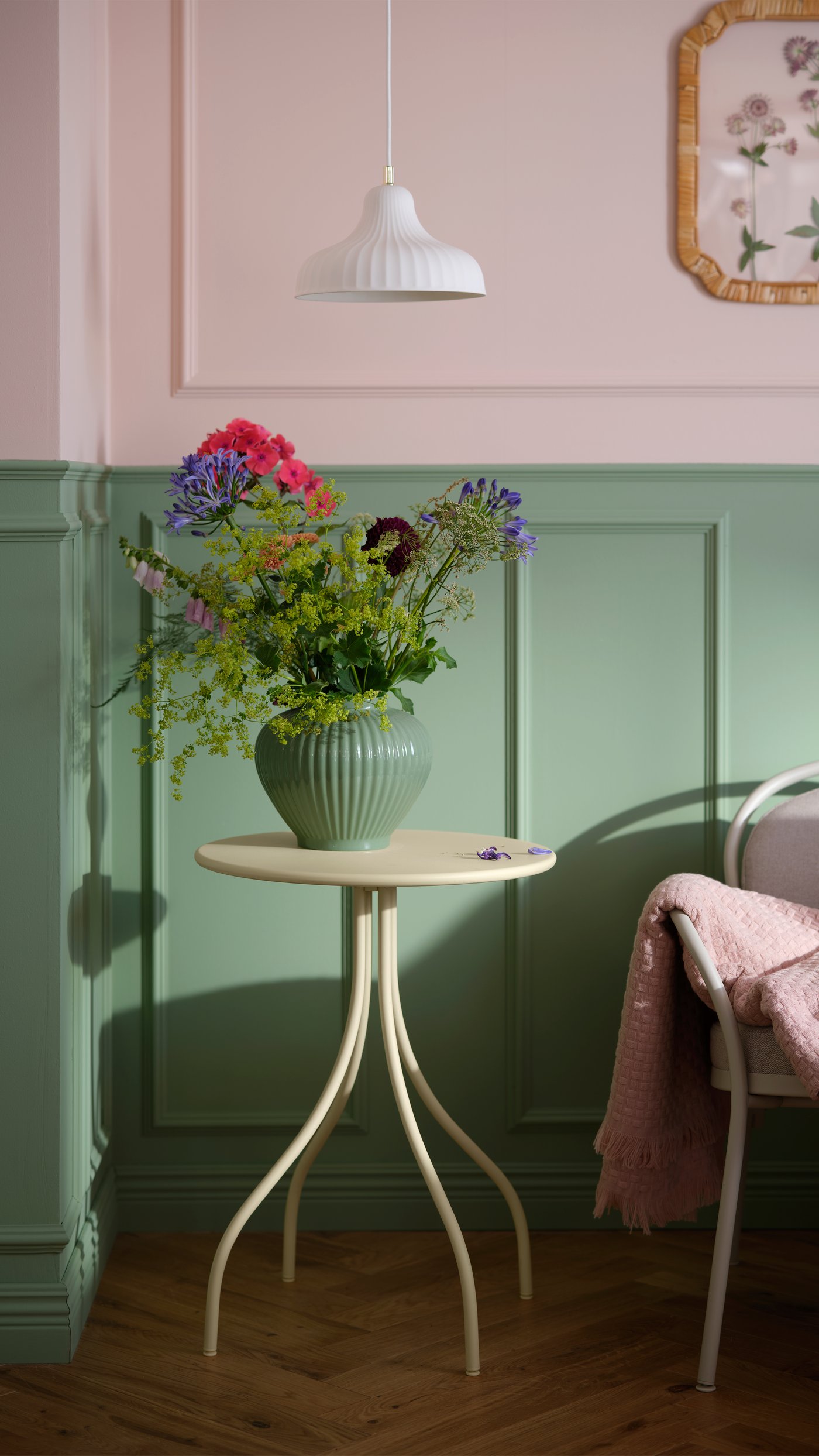 Litet runt bord i ljus trä med en grön vas fylld med färgglada blommor. Bordet står mot en vägg med gröna paneler och rosa övre del. Ovanför hänger en vit taklampa, och till höger syns en stol med rosa pläd.