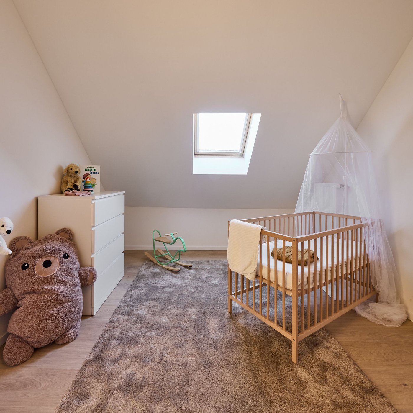 Gezellige babykamer onder schuin dak met houten wieg, commode, knuffels en dakraam met natuurlijk licht.