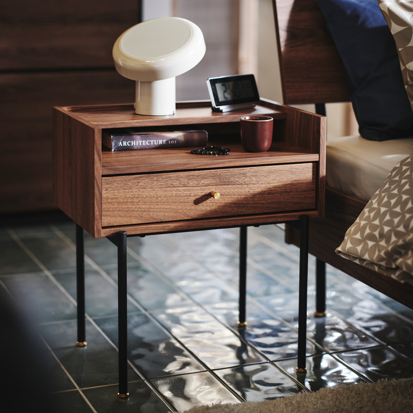 Close-up of RÅDMANSÖ bedside table in a walnut colour with some knick-knacks and BLOMPRAKT Bluetooth speaker lamp on top.