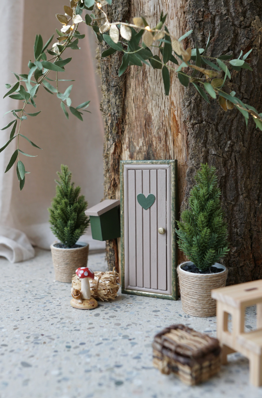 Kleine Wichteltür mit Herz an einem Baumstamm, daneben zwei Mini-Tannen in Töpfen, ein Briefkasten und weihnachtliche Dekoration in gemütlicher Adventsstimmung.