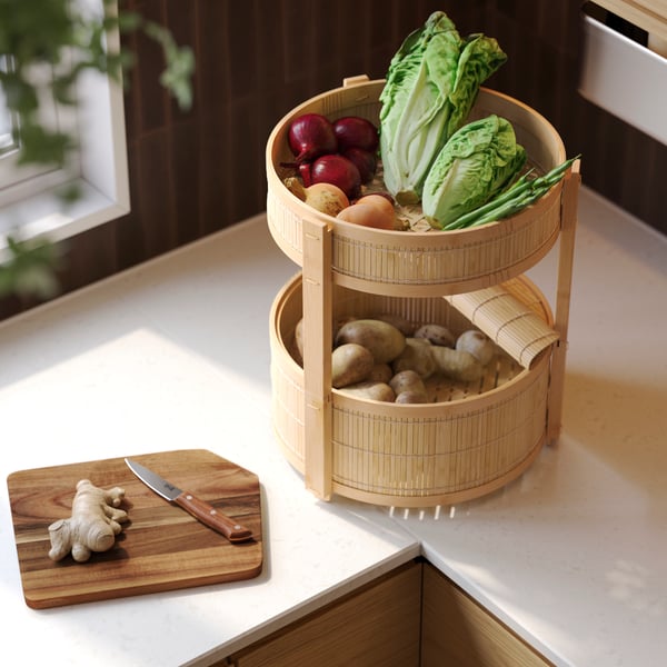 A worktop with a KANELMOTT vegetable and fruit basket in rattan and bamboo containing vegetables, a chopping board.