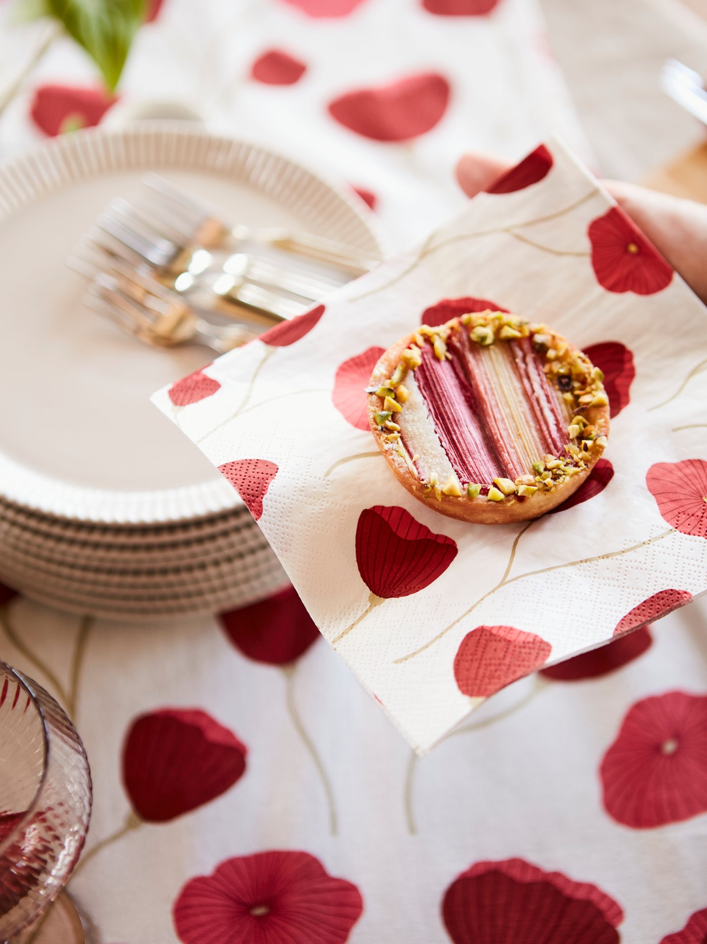 An ANLEDNING paper napkin with a poppy pattern holding a mazarin cake, with a stack of plates and poppy tablecloth behind.