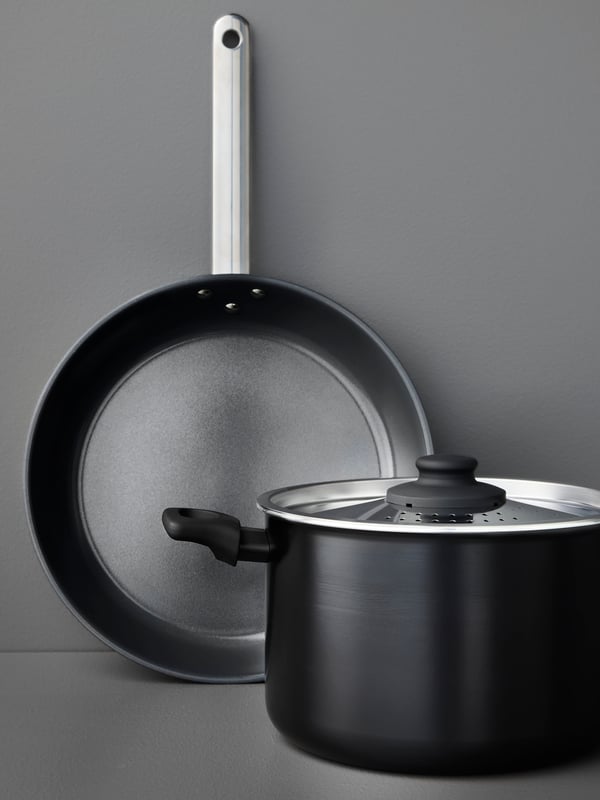 A black HEMLAGAD frying pan and a HEMLAGAD pot with lid, both with non-stick coating, are placed on a grey surface.