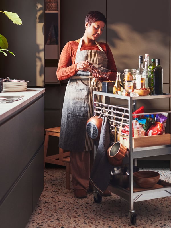 Una persona accanto a un carrello da cucina KUNGSFORS in acciaio inossidabile con cibi e prodotti per cucinare, in una cucina con pareti grigio scuro.