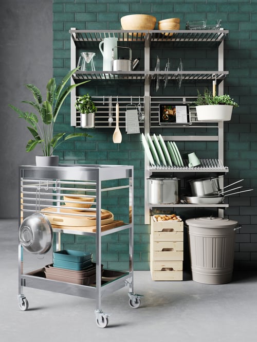 A steel KUNGSFORS kitchen trolley holds serving trays, oven dishes and a plant. It is by wall shelves full of kitchen items.