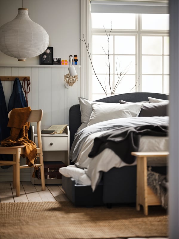 A room with a large bed by a window, grey BLÅVINDA bed linen, a white HAUGA bedside table and a white RISBYN pendant lamp.