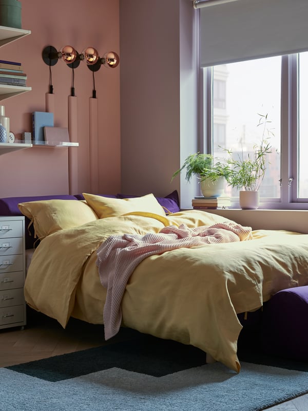 FLOTTEBO sofa-bed with yellow covers, in a room with storage cabinets, beside a window with plants in the sill.
