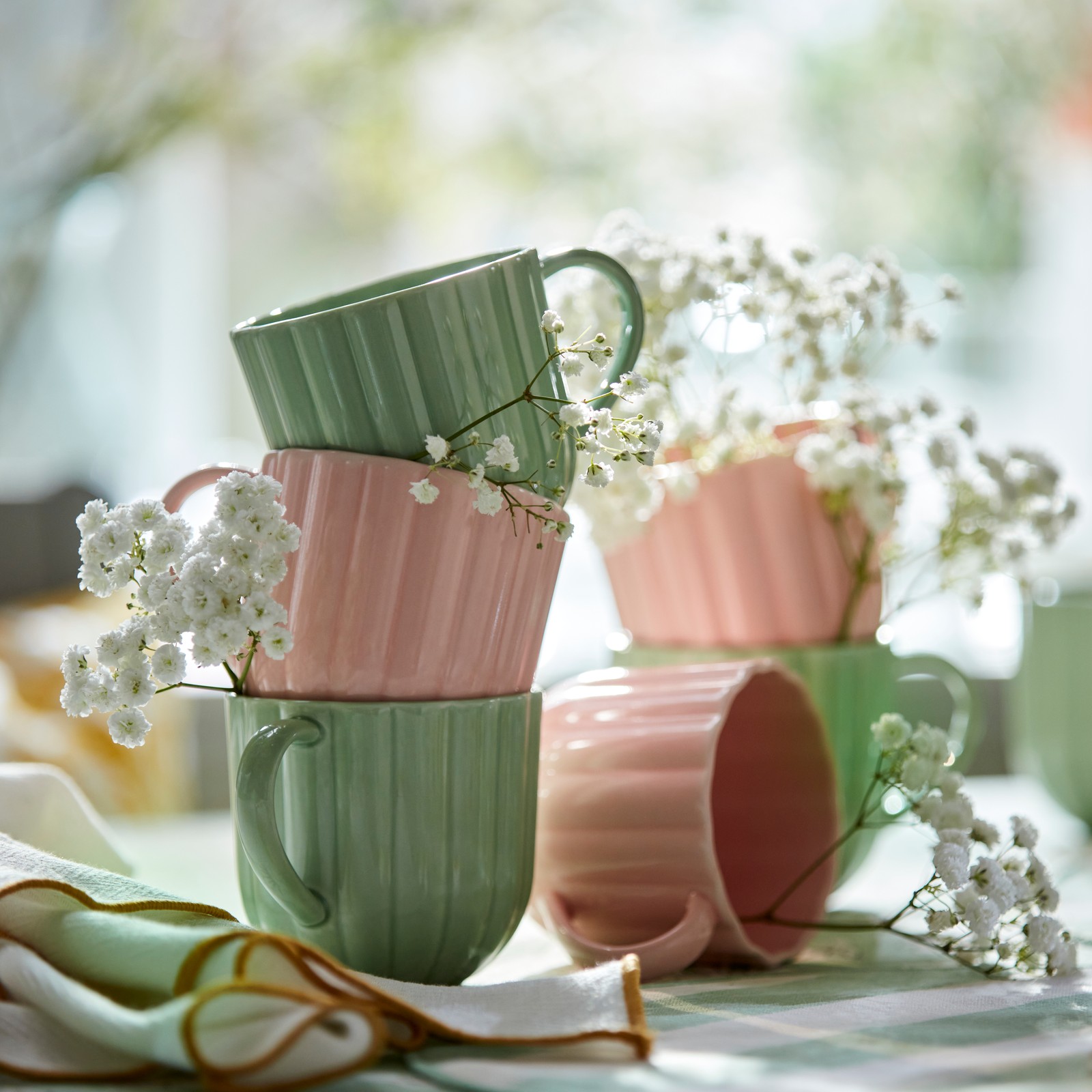Some light green and pink SMÖRFISK mugs surrounded by delicate white flowers create a fresh and vibrant atmosphere.