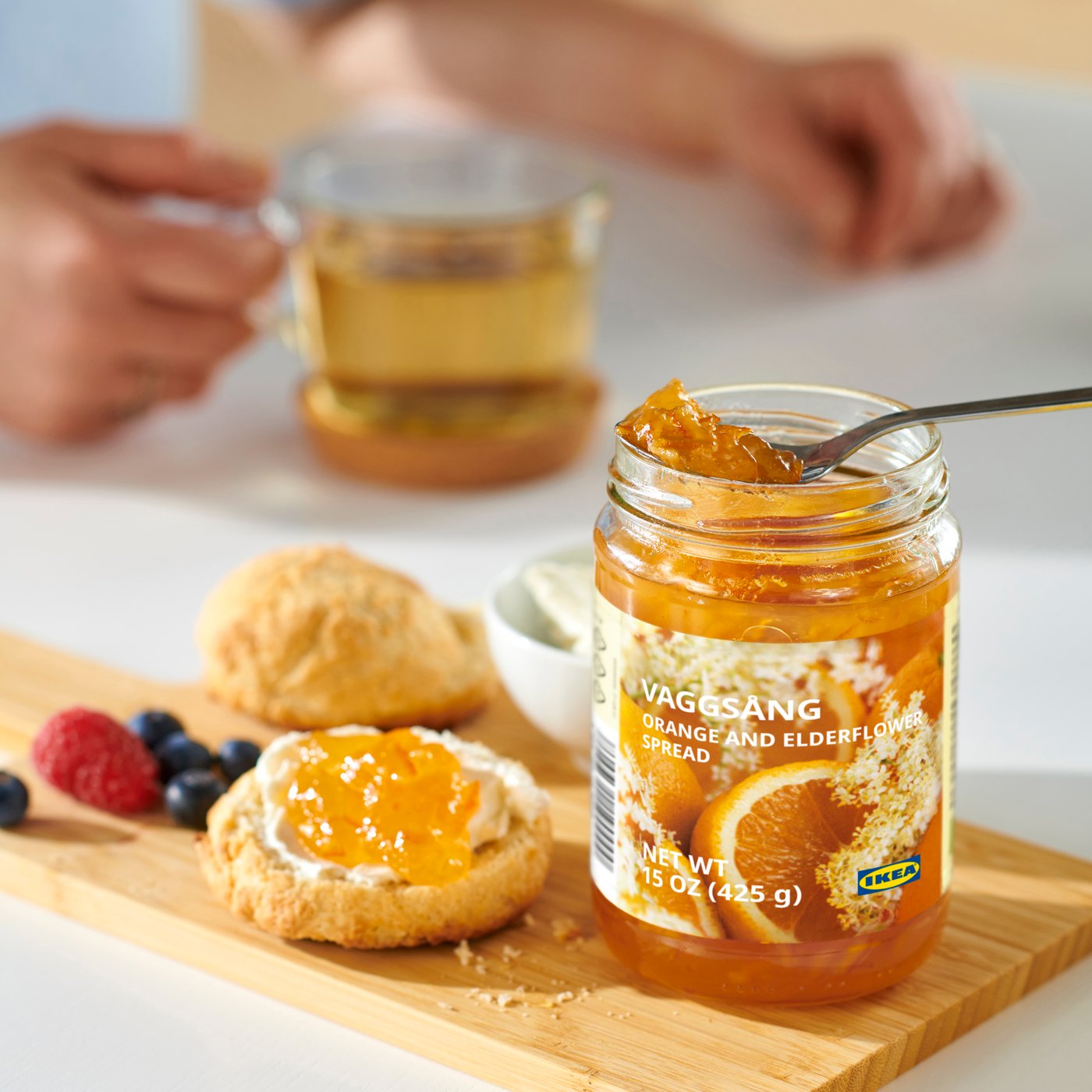 On a wooden board stands an open jar of VAGGSÅNG orange and elderberry jam, with a spoon inside, as well as berries and a round crispbread topped with the jam. In the background, a person behind a table is holding a transparent mug filled with tea.