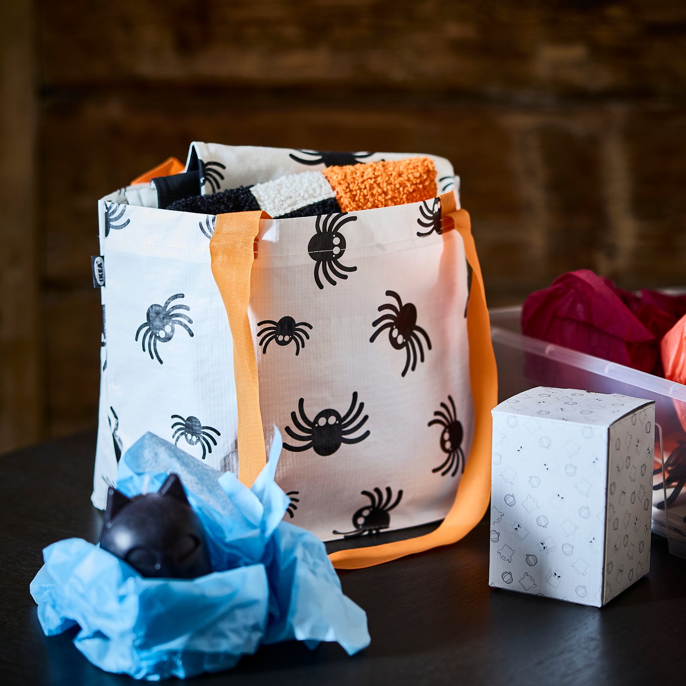 A white spider-patterned KUSTFYR carrier bag on a tabletop beside a KUSTFYR black cat-shaped candle wrapped in tissue paper.