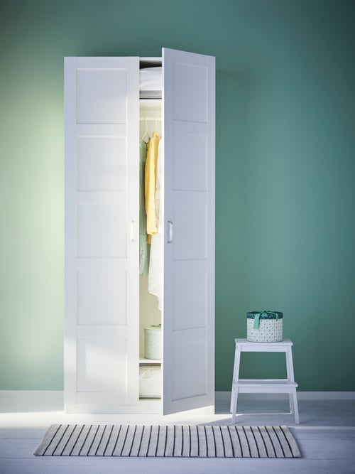 Bedroom setting with green wall with a white wardrobe with one door open, a white step stool, and a flatwoven rug.