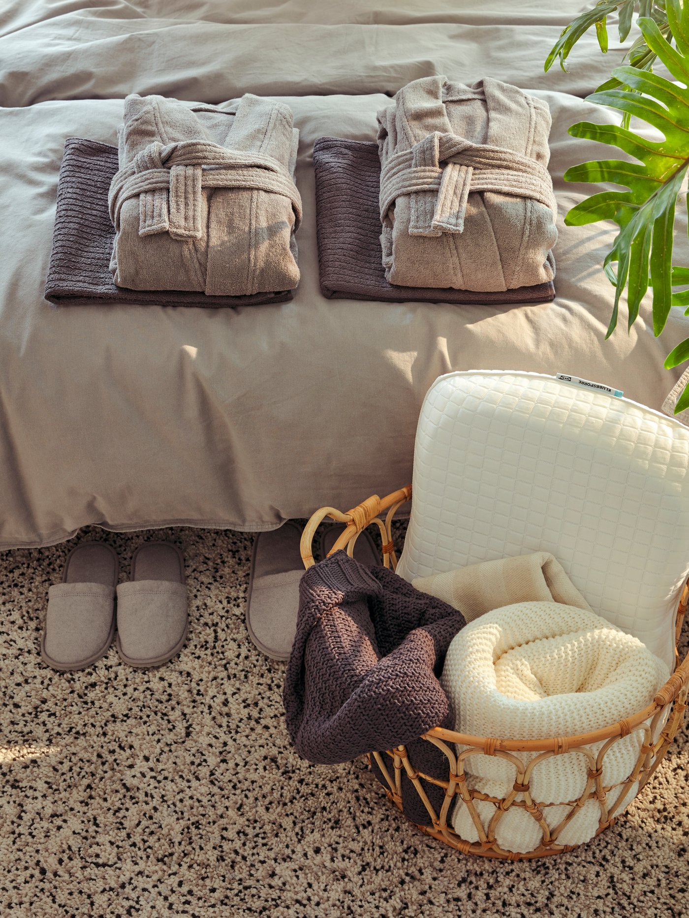 ROCKÅN bathrobe on bed with TÅSJÖN slippers on carpet and KLUBBSPORRE pillow in SNIDAD basket.