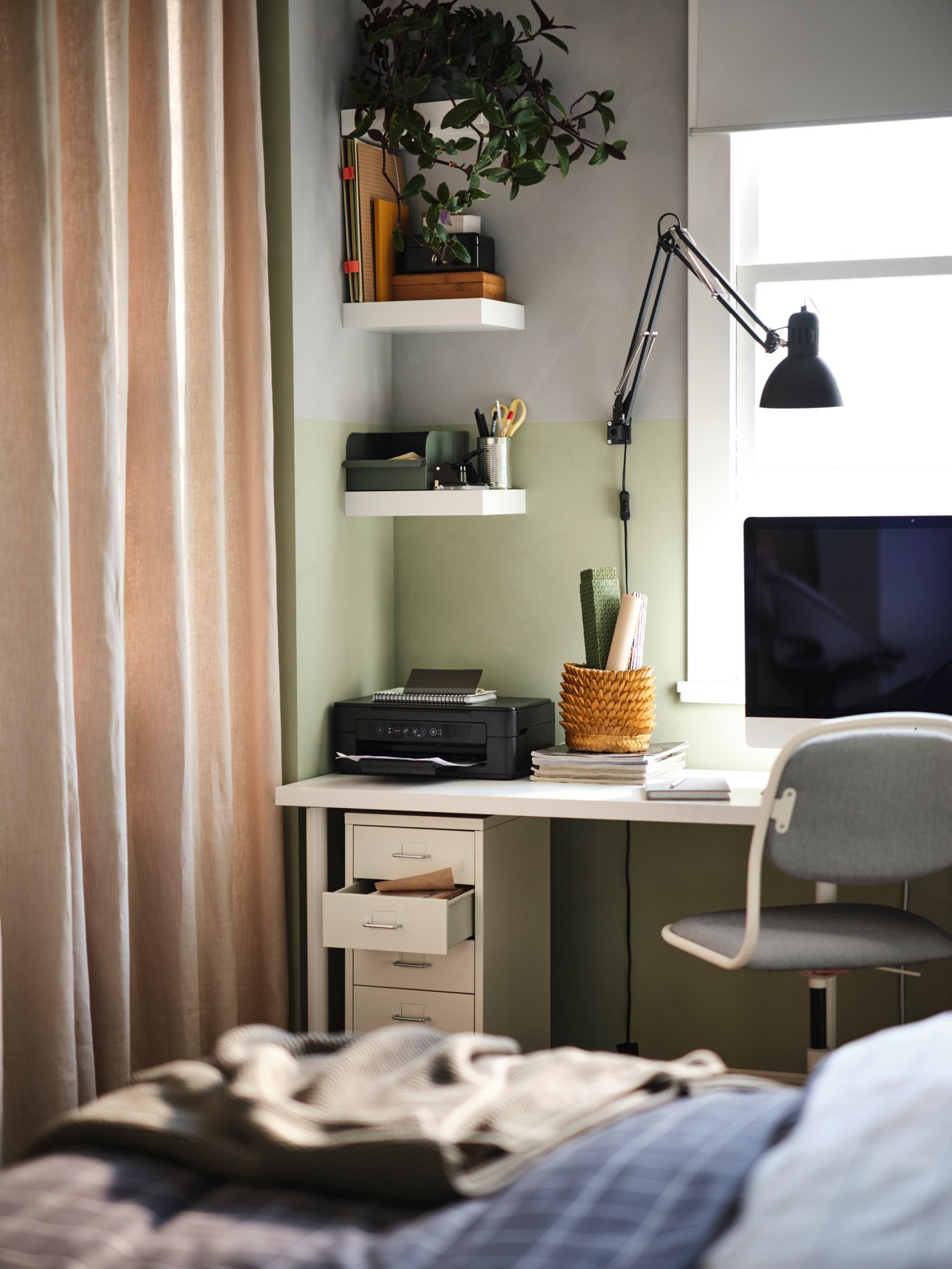 Dormitorio con una cajonera blanca con ruedas, estantes de pared, un escritorio con un monitor y una lámpara de trabajo, y una silla giratoria gris.