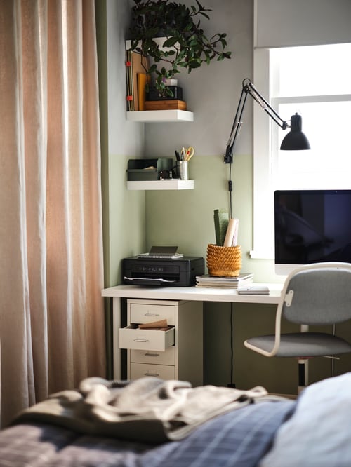Bedroom with a white drawer unit on castors, wall shelves, a desk with a monitor and a work lamp, and a grey swivel chair.