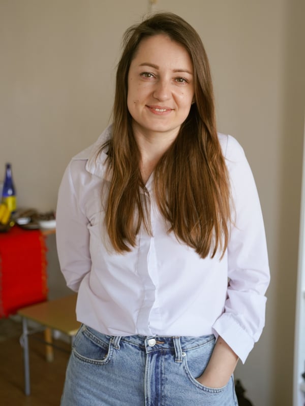 A girl stands indoors and looks into the camera in a white shirt and brown loose hair.