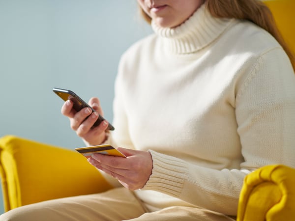 Una donna seduta su una poltrona gialla STRANDMON mentre sta facendo un pagamento con la carta tramite smartphone.