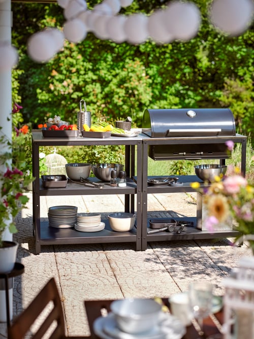 A GRILLSKÄR barbecue on a sunny terrace with vegetables in bowls, cooking utensils and serving bowls on the shelves.