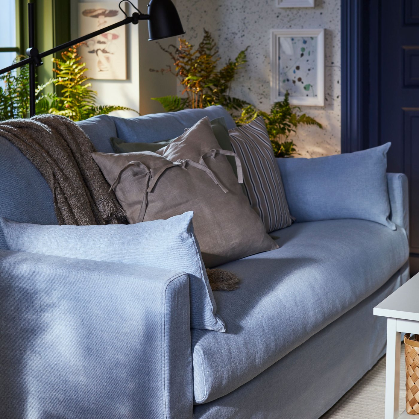 Es ist ein hellblaues HYLTARP 2er-Sofa mit einem Plaid und zwei zusätzlichen Kissen zu sehen. Ein Kissen besitzt einen beigefarbenen AINA Kissenbezug.