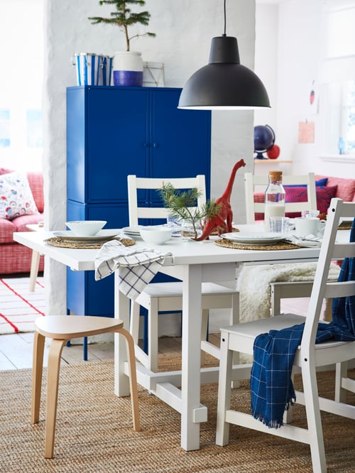 Dining area with table, chairs, stool, flat woven rug, throw, carafe, mugs, glass, pendant lamp, storage unit, toys, bowls.
