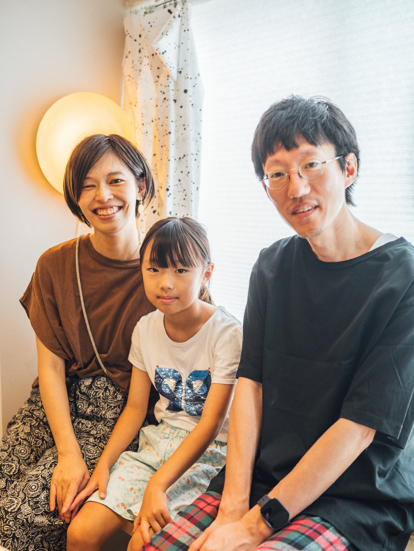 明るい部屋で、丸い照明の前に大人2人と子ども1人が並んで腰掛け、カジュアルな服装でこちらを向いている。