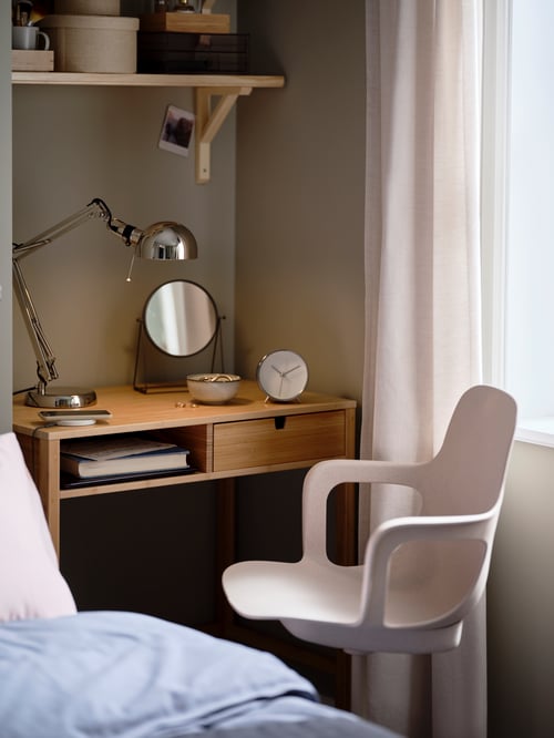 Modern workspace that features a dressing table, a table mirror, a work lamp, an alarm clock, a bowl, and a swivel chair.