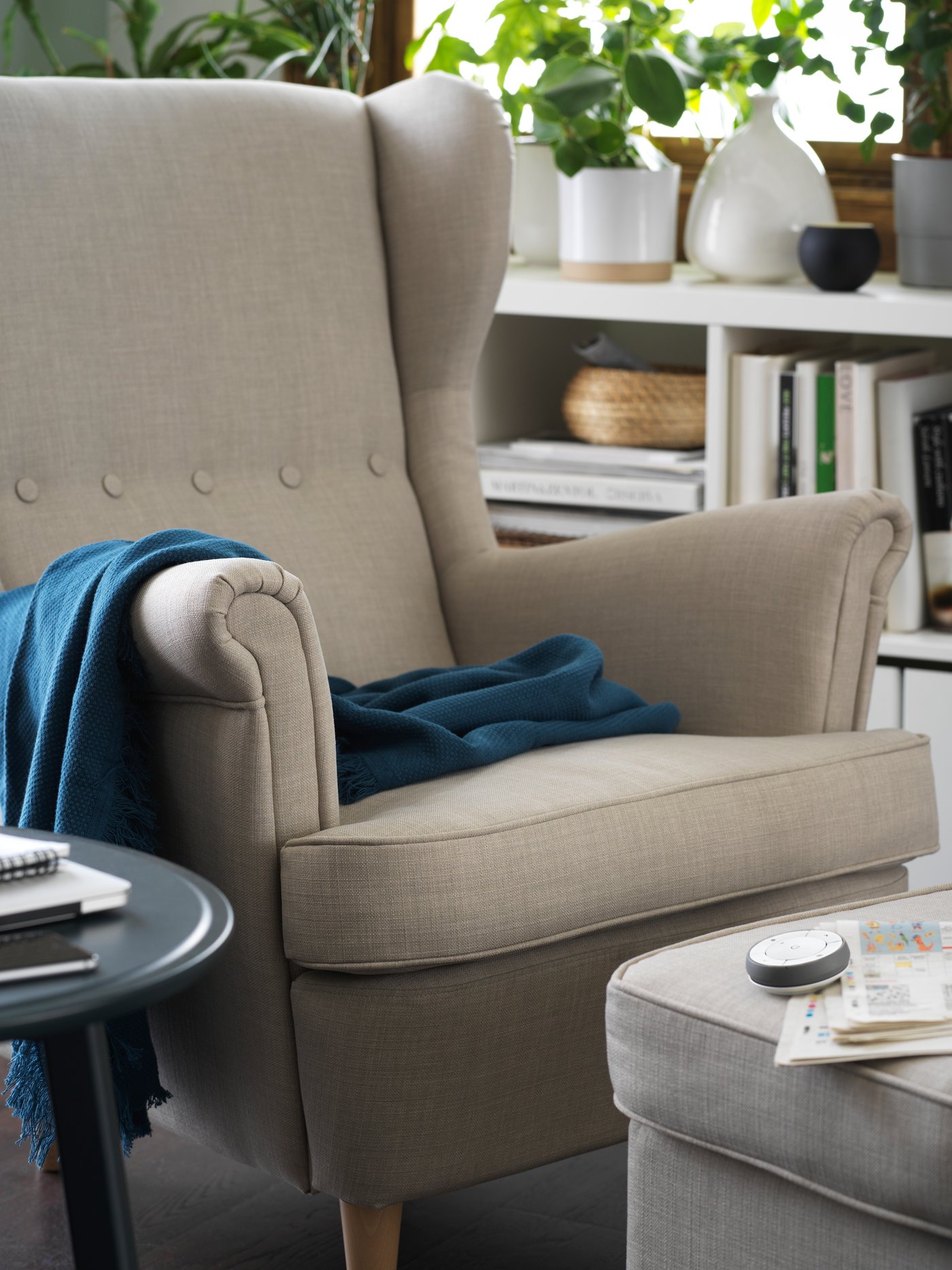 TRÅDFRI remote control device and a newspaper on a STRANDMON footstool in front of a STRANDMON armchair, both light beige.