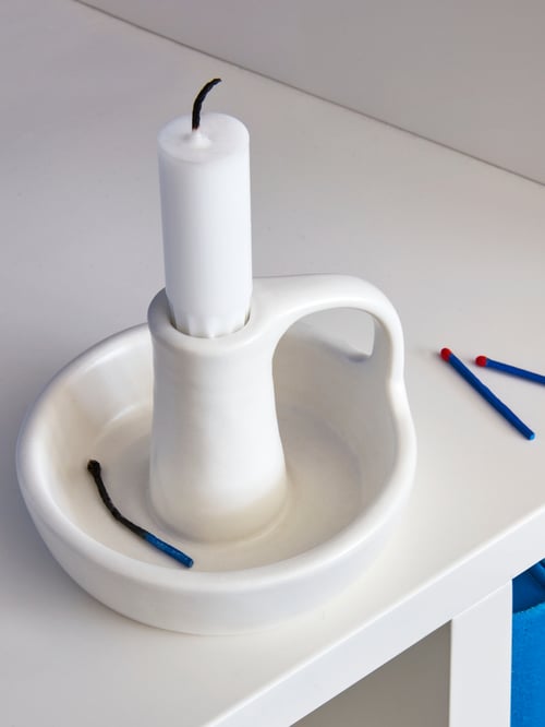 White ceramic GODTAGBAR candlestick holder with tapered candle sits on a white table; surrounded by several wooden matches.
