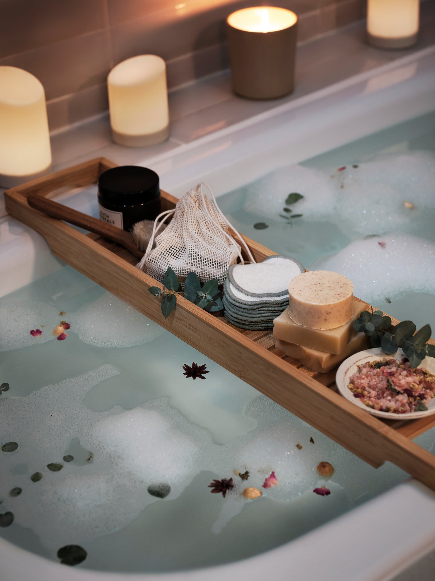 Tab mandi dengan lilin wangi dan rak mandi HAVERN buluh yang mempunyai pad pembersih boleh guna semula ÅKERRÄTTIKA dan item-item lain.