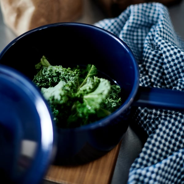 A blue VARDAGEN enamelled steel saucepan with kale inside, next to a blue and white check kitchen towel.