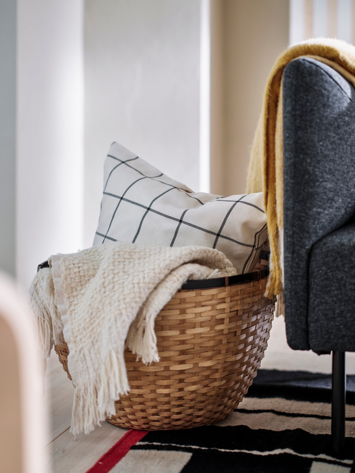 A LÖVRÄFSA basket containing a HUMLEMOTT throw and a cushion with a SPIKKLUBBA cushion cover stands on a RASTPLATS rug.