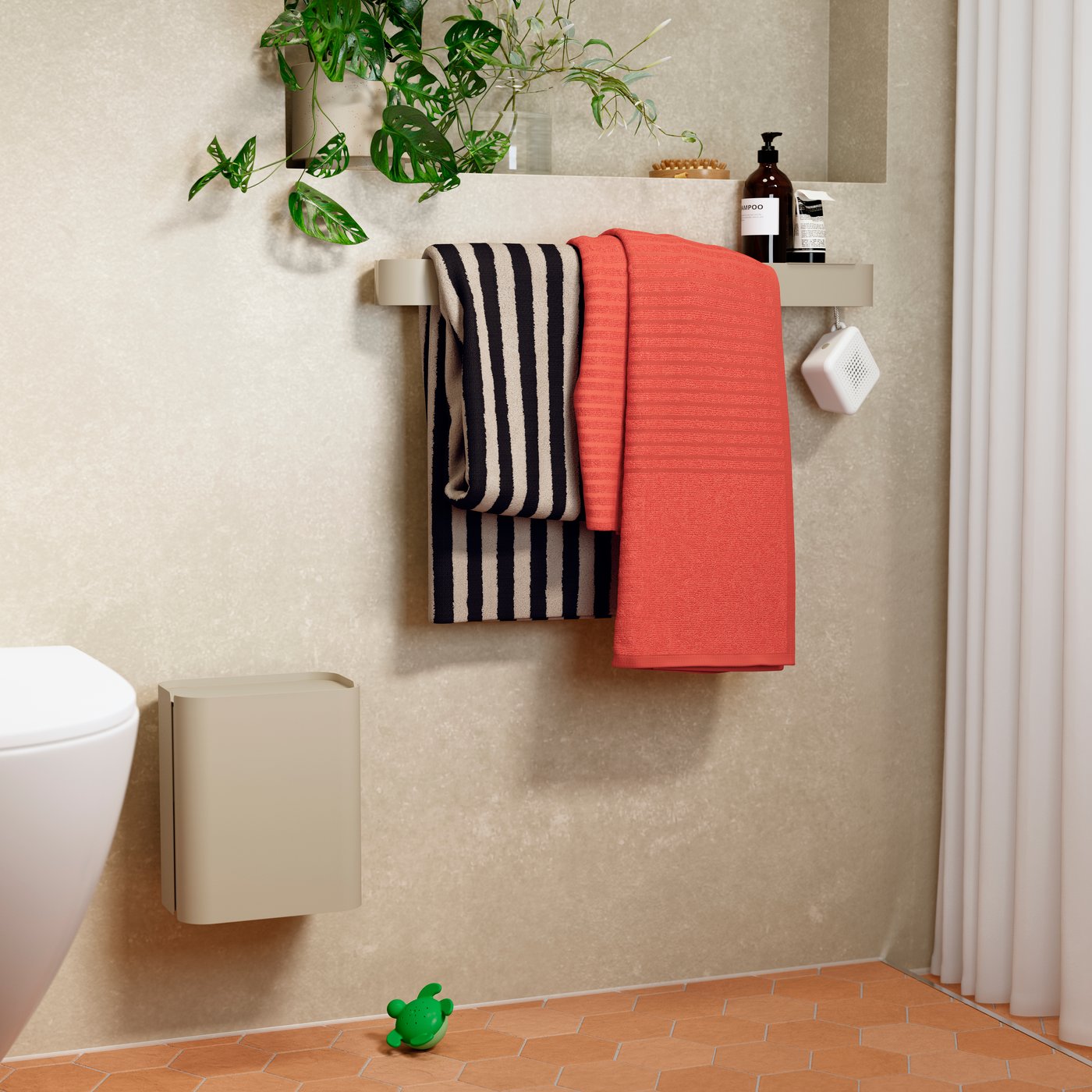 IKEA-badkamerhoek in zachte beige tint met een ENHET-wandplank en een BROGRUND-zeepdispenser, een zwart‑wit gestreepte en een koraalrode handdoek aan de plank, een hangende kamerplant erboven, een klein groen rubber kikker‑speeltje op terracotta tegels en een wandgemonteerde beige prullenbak.