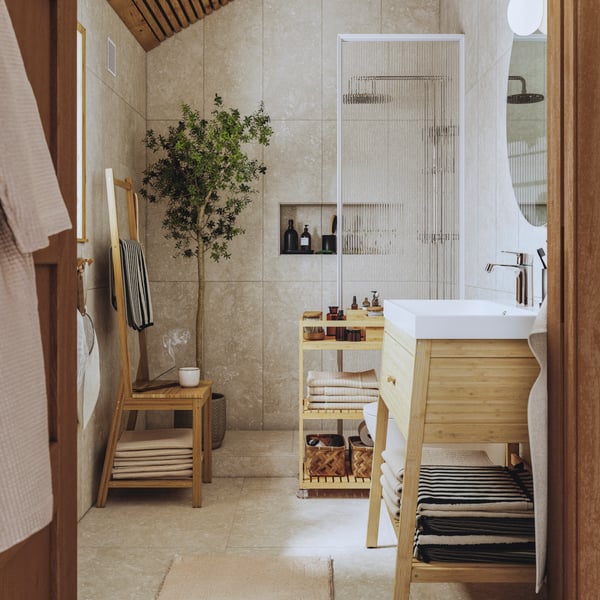 A sunlit bathroom with RÅGRUND trolley, towel rack chair and wash stand in natural bamboo. A large plant is in the corner.