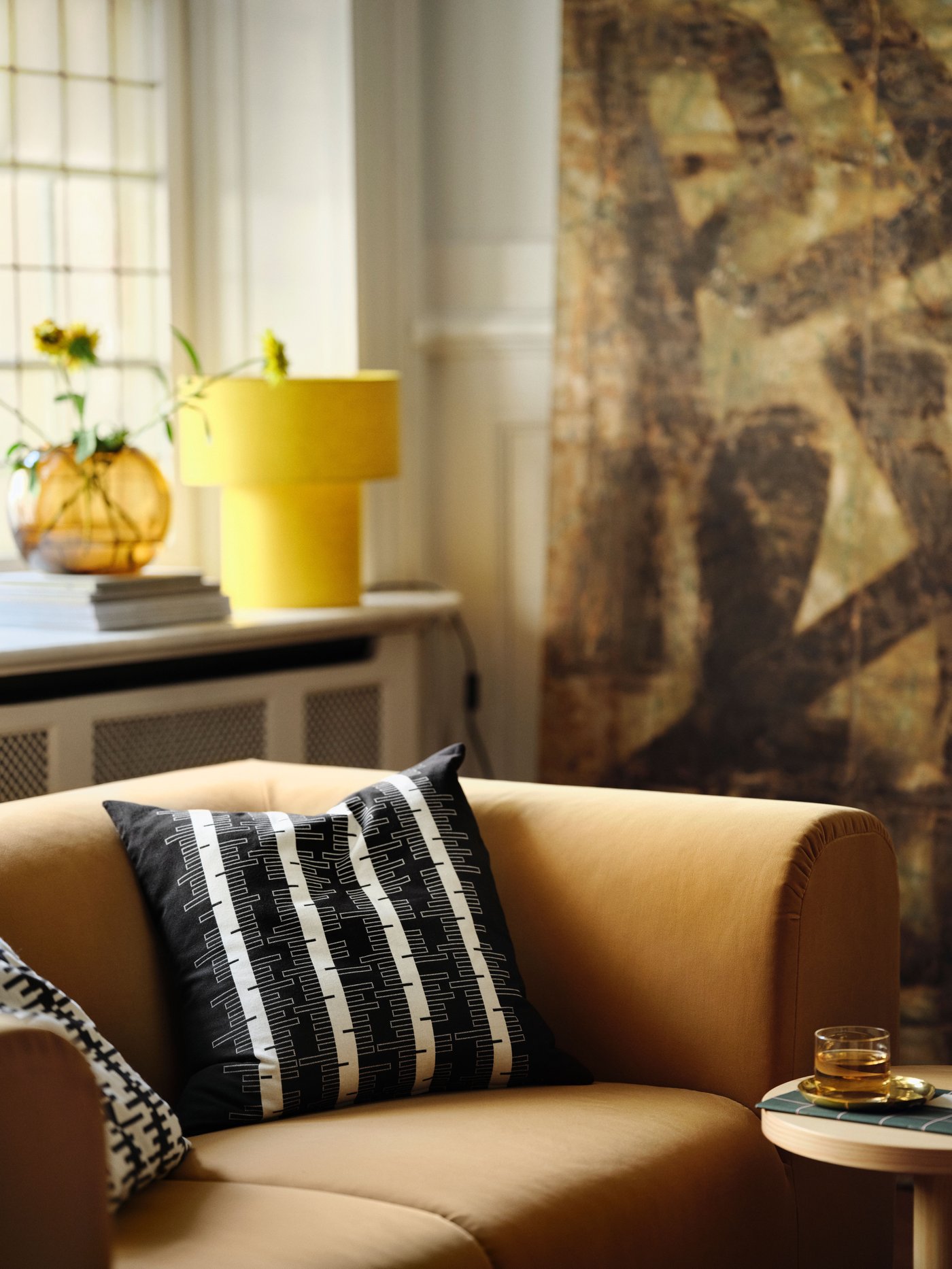 A cushion with a black and off-white STOCKHOLM 2025 cushion cover resting in a beige 2-seat sofa in front of a window.