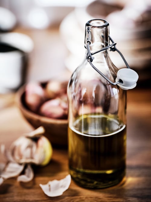 A glass bottle with a stopper on a wooden surface, half-filled with olive oil, beside a bowl of garlic bulbs.