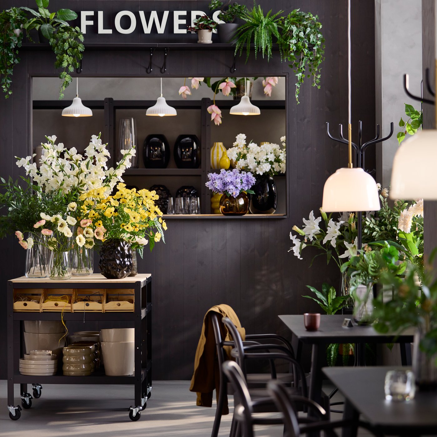 Café enviroment with tables, chairs and a black BROR trolley made from black/pine plywood, filled with flowers and greenery.