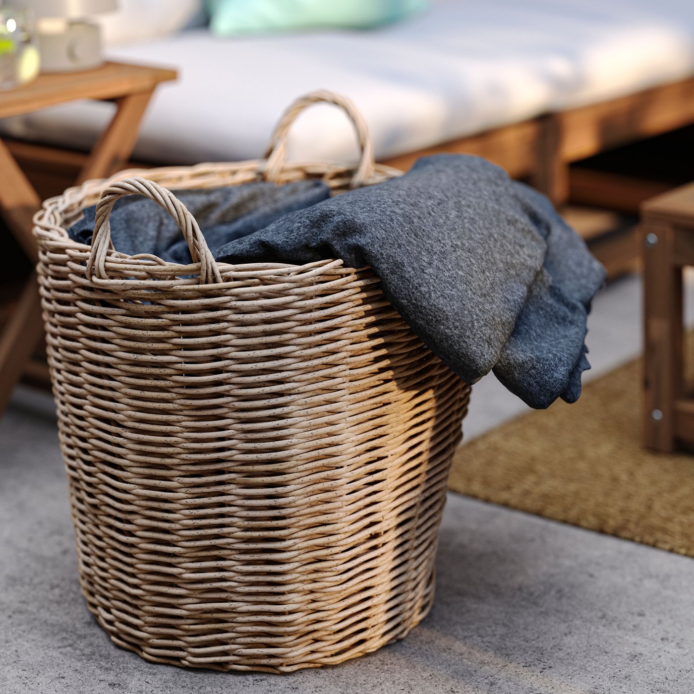 A lounging zone on a patio with a light grey/brown in/outdoor FLODBJÖRK plant pot in rattan working as storage for blankets.