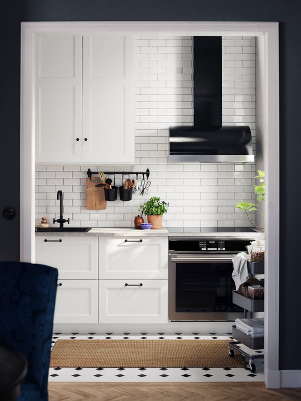 A kitchen with AXSTAD matte white kitchen fronts with black handles, a black tap, a cart, against a tiled wall.