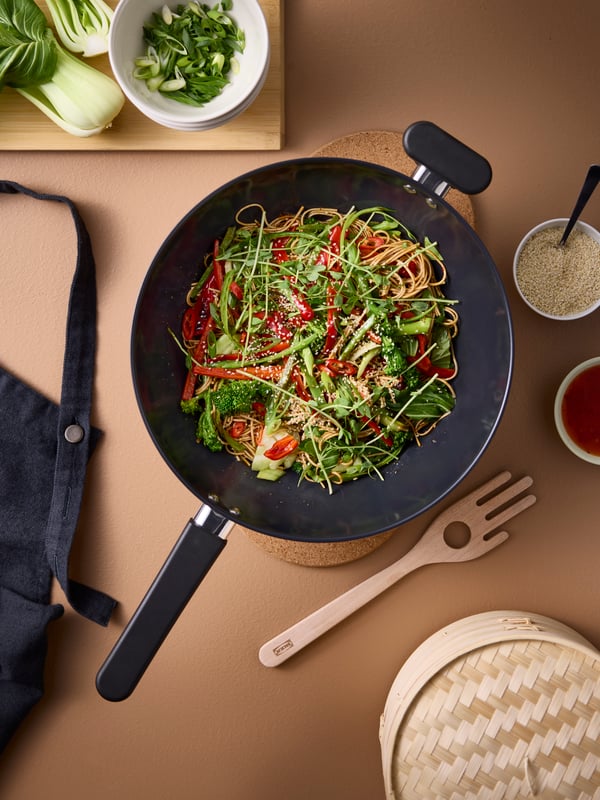 A hand is using a wooden spoon to serve a vegetable and noodle dish out of a MIDDAGMAT wok into two black bowls next to it.