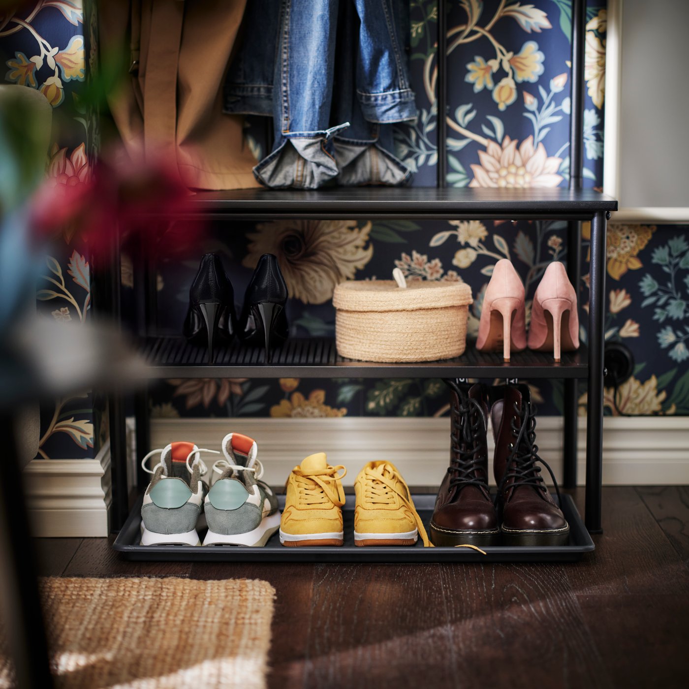 Auf der Bank eines schwarzen NIPÅSEN Garderobenständers mit Bank/Schuhaufbewahrung stehen vier Paar Schuhe, ein Paar Stiefel und ein Korb.